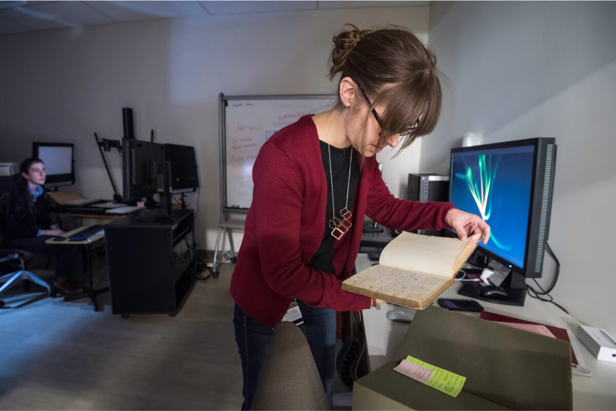 librarian in a digital lab surrounded by computer screens holds an old book.