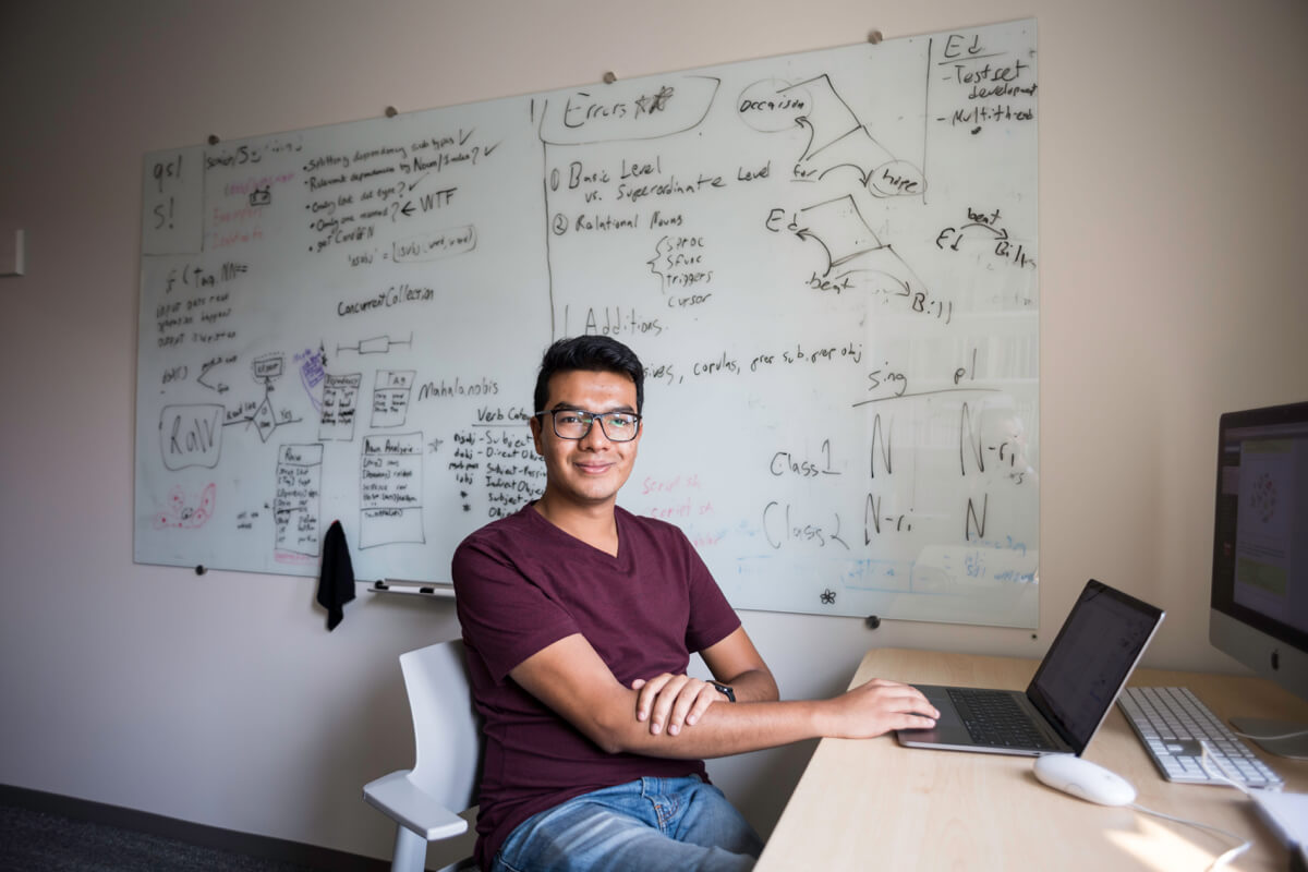 A person sitting at a desk with a laptop, in front of a whiteboard covered in notes and diagrams.