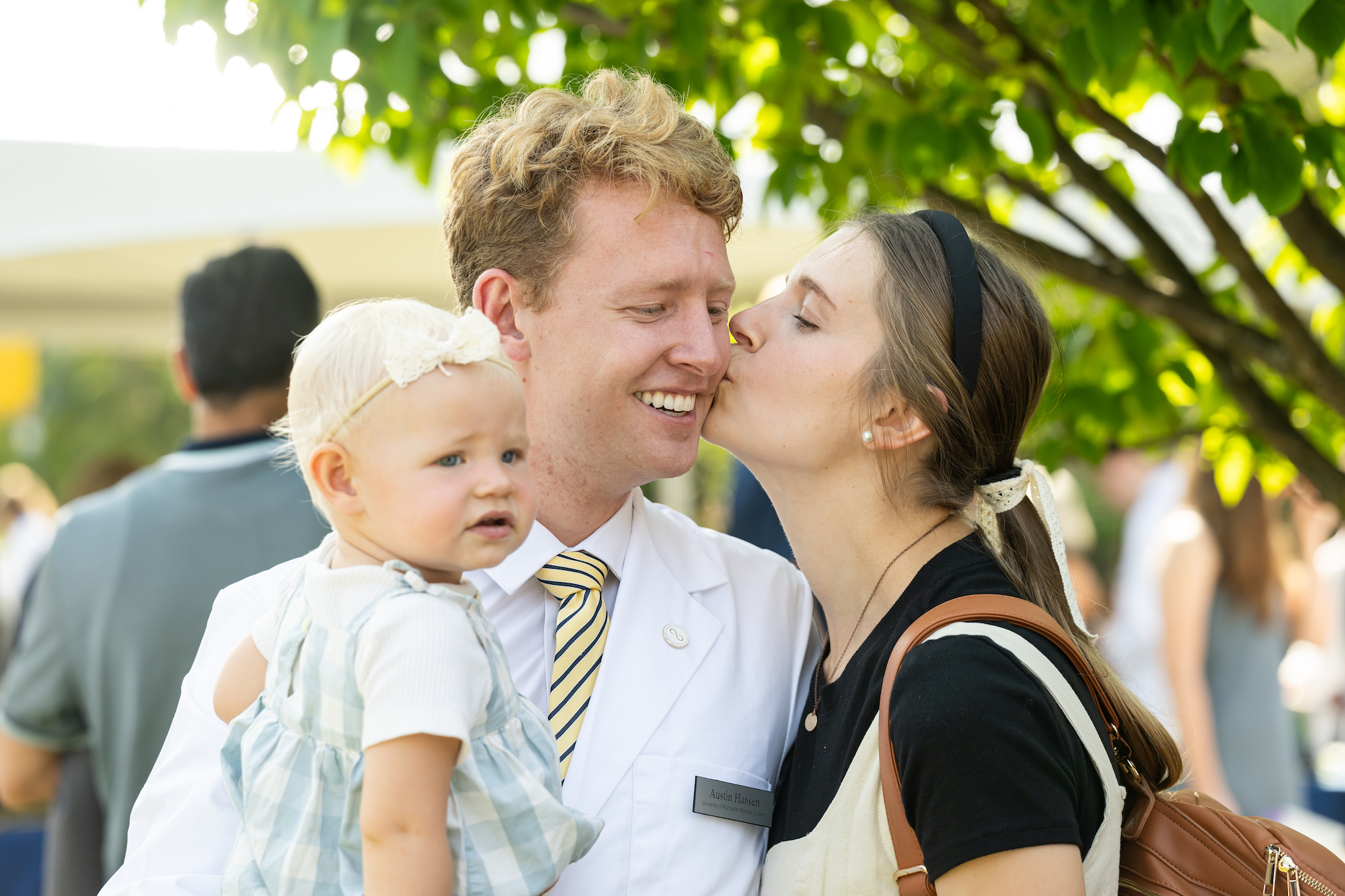 A man in a white medical coat gets a kiss on the cheek from his wife, while holding his infant daughter