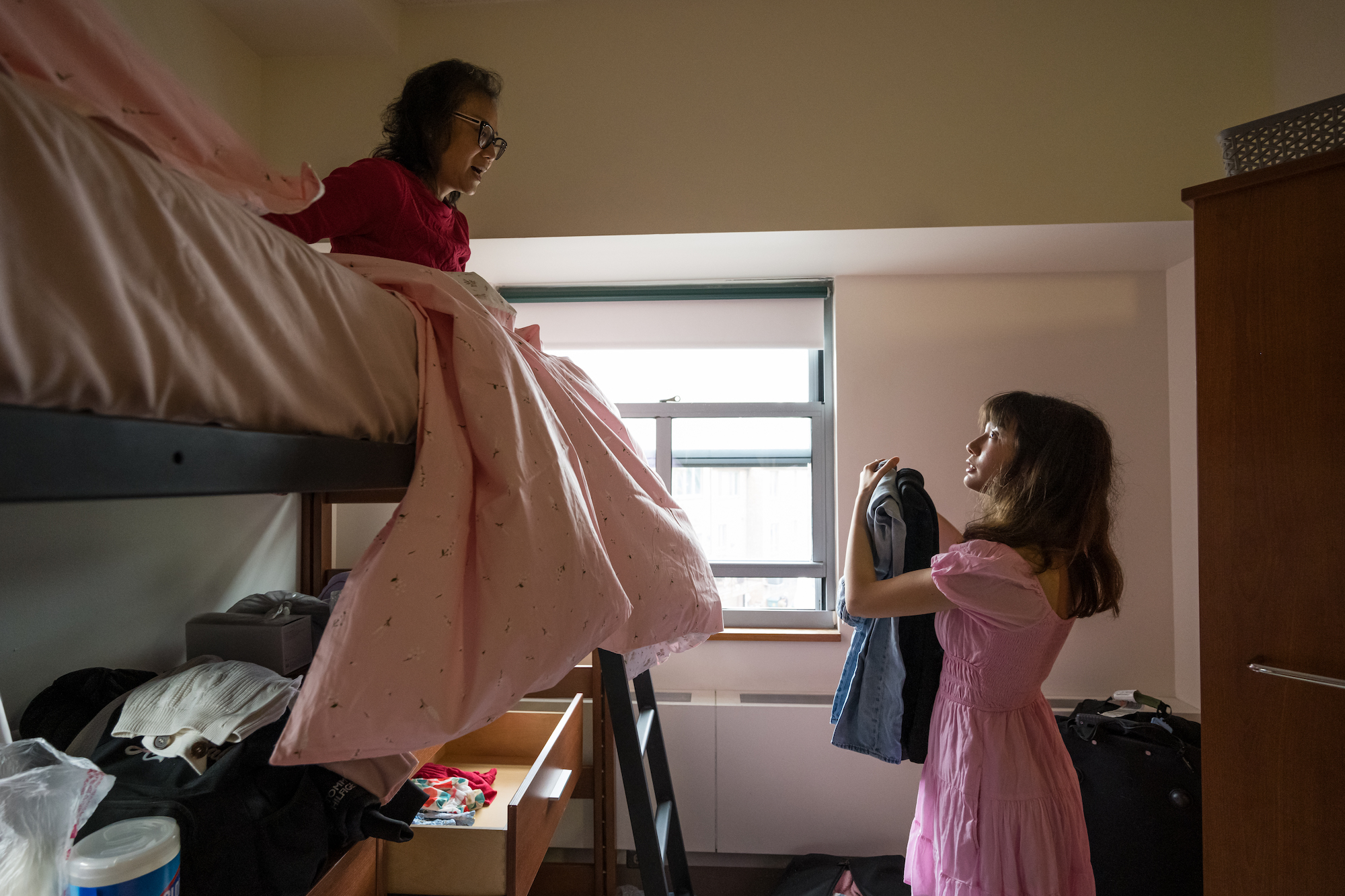A mother atop a bunk bed in a dorm room, assisting her daughter with moving in
