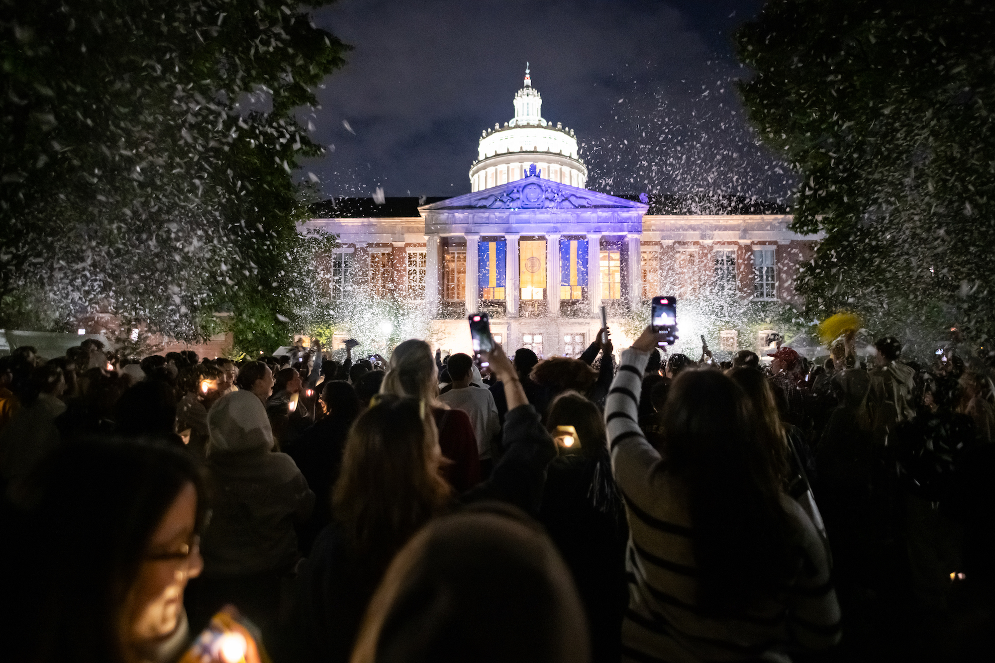 Exterior of Rush Rhees Library, showing students by candlelight and surrounded by confetti
