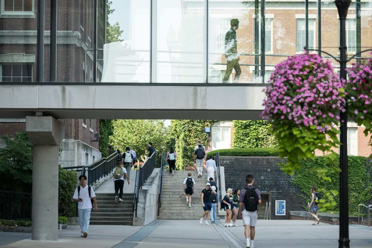 College students walking on a campus under a glass pedestrian bridge with a staircase and brick buildings in the background.
