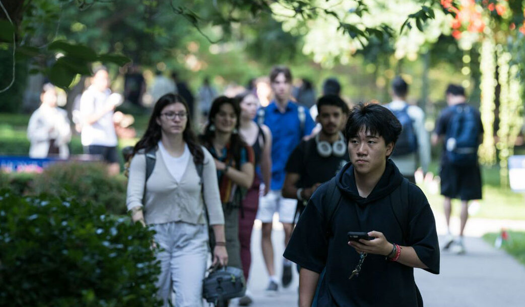 A group of people walk on a paved pathway in a park or garden, surrounded by green trees and bushes.