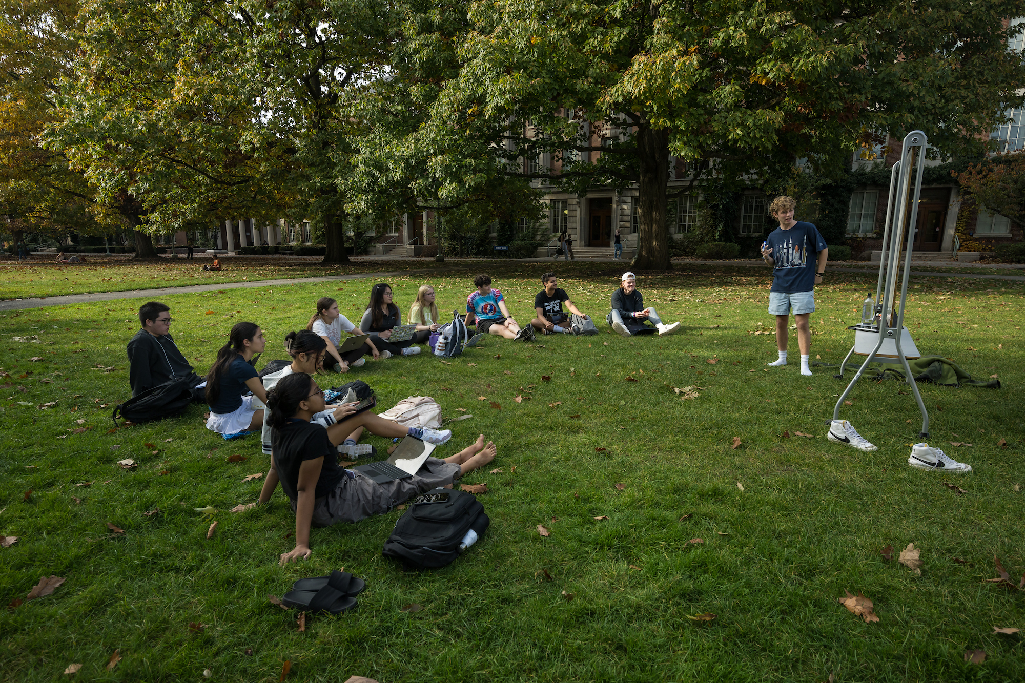 students sit outside on the grass in a semi-circle while another student, standing, leads the class
