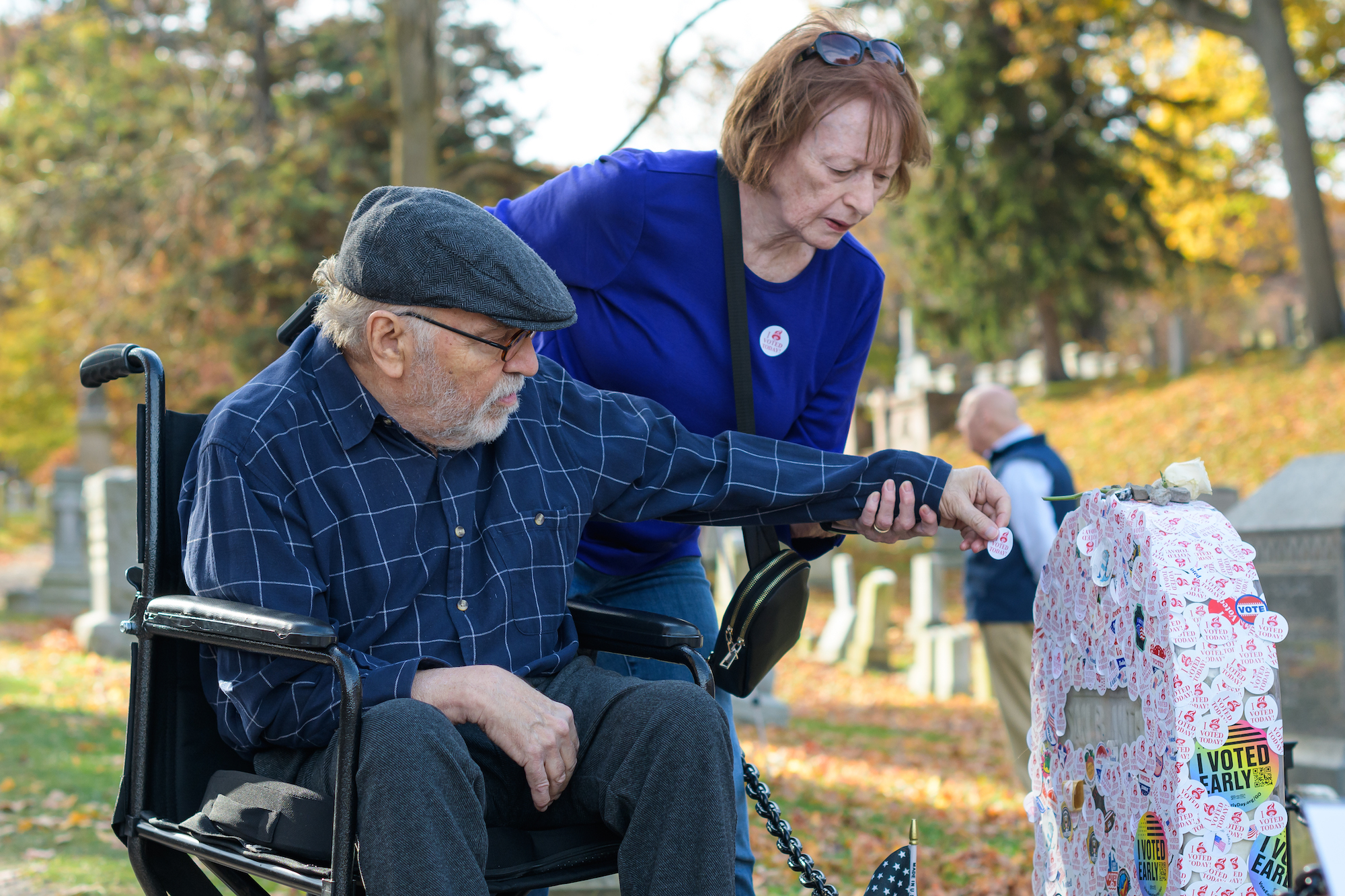 Woman assists man in wheelchair in putting I Voted sticked on the headstone of Susan B. Anthony