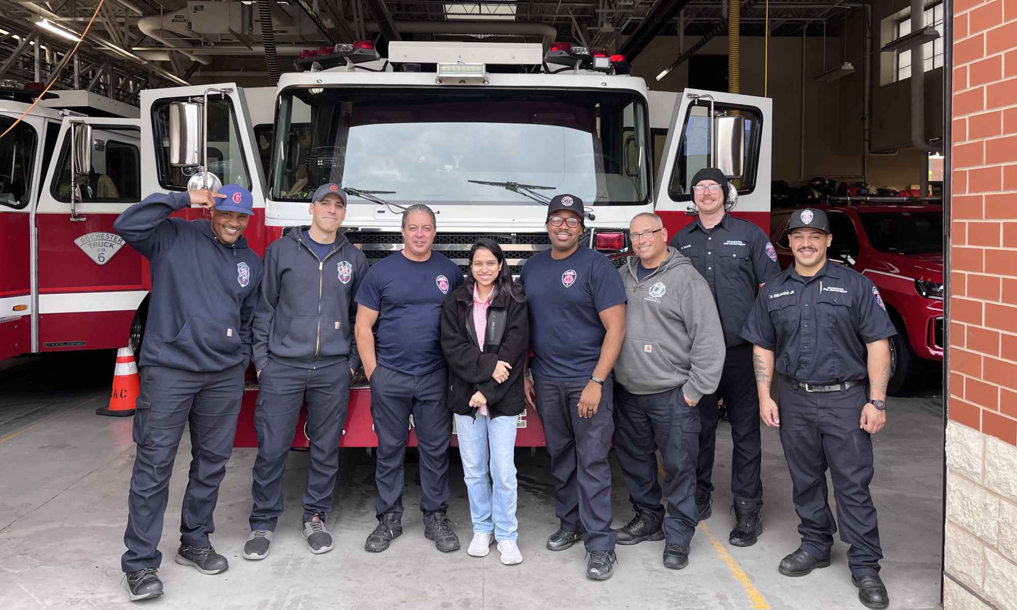 A University of Rochester data science student flanked by seven members of the Rochester Fire Department with a fire truck and equipment in the background.