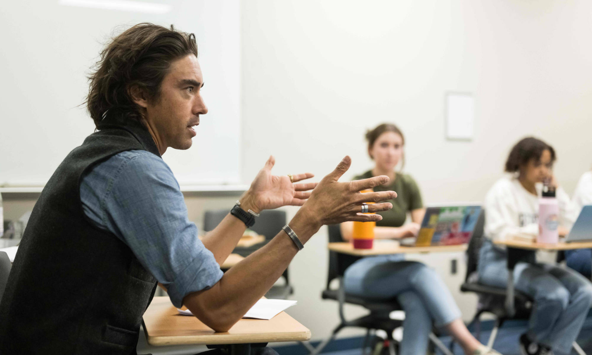 Jack Downey seated at a desk in a classroom teaching with two students seated with their laptops out of focus in the background.
